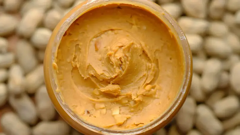 An open jar of creamy peanut butter surrounded by peanuts